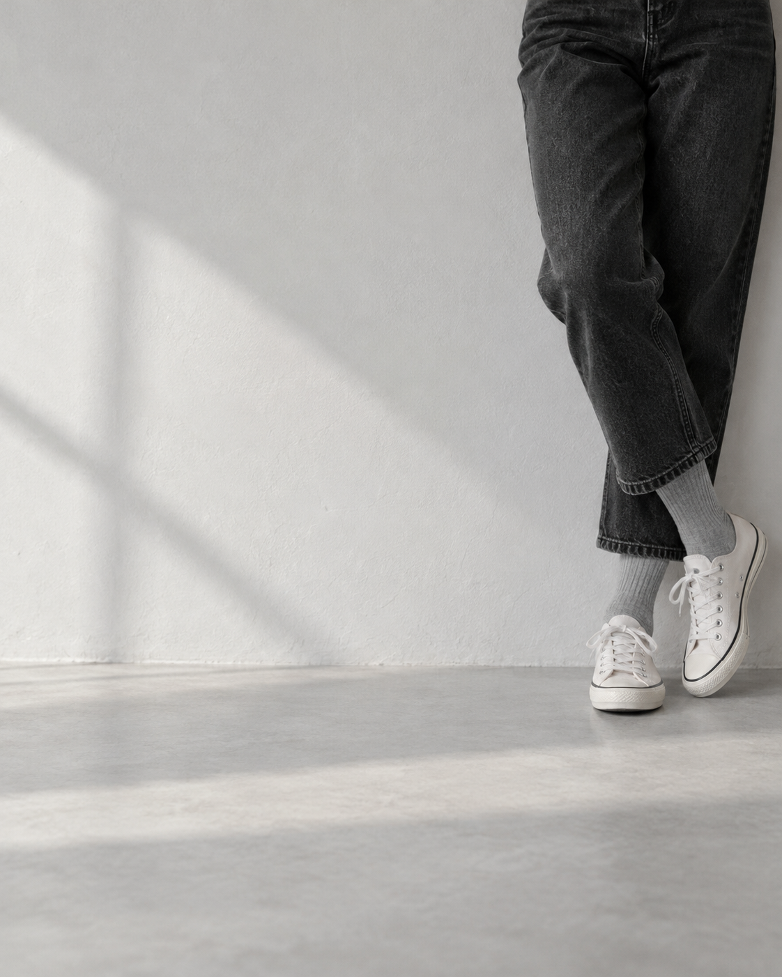 Leaning against a pale wall in black denim, white canvas sneakers, and Sleet Grey crew socks